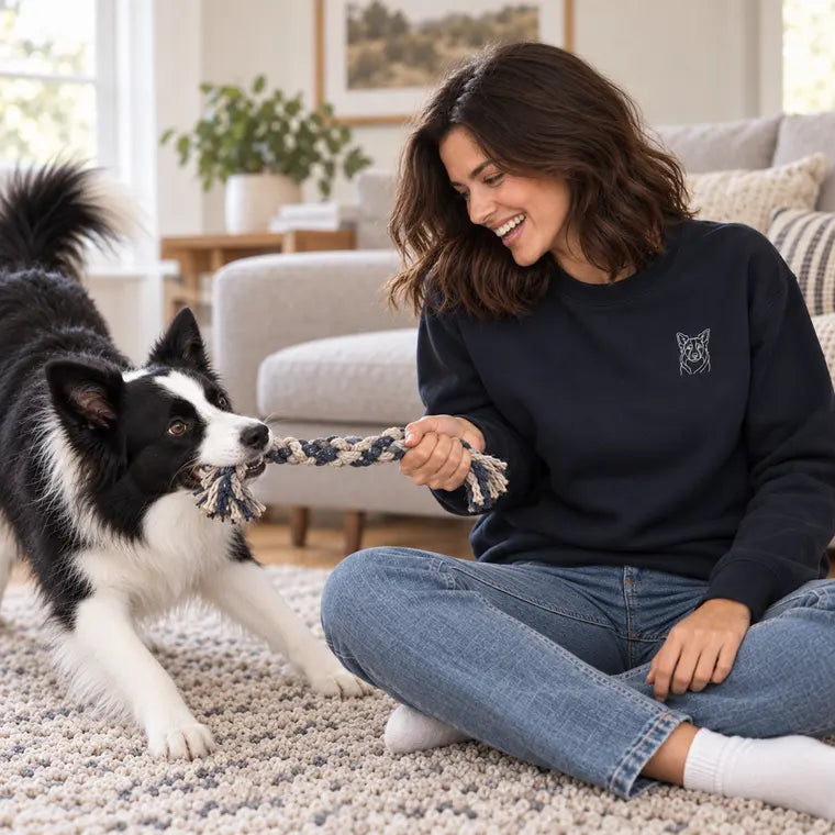 A woman in a navy sweatshirt embroidered with her border collie's face, playing tug with him on the rug.